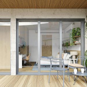 A perspective looking from an outdoor wooden terrace through large glass sliding doors into a modern bedroom. The balcony area features wooden decking, two grey metal chairs, a small round table with coffee mugs, and a wall covered in climbing green ivy. Inside the room, a bed with pillows, a desk workspace, and shelving are visible. The architecture combines raw concrete walls with warm wood elements, and reflections in the glass suggest an urban setting.